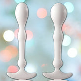 Two white, elongated objects with a bulbous end and a flat base stand vertically against a blurred background of colorful bokeh lights, reminiscent of the Aneros Peridise Anal Stimulators (2-Pack) by Aneros.